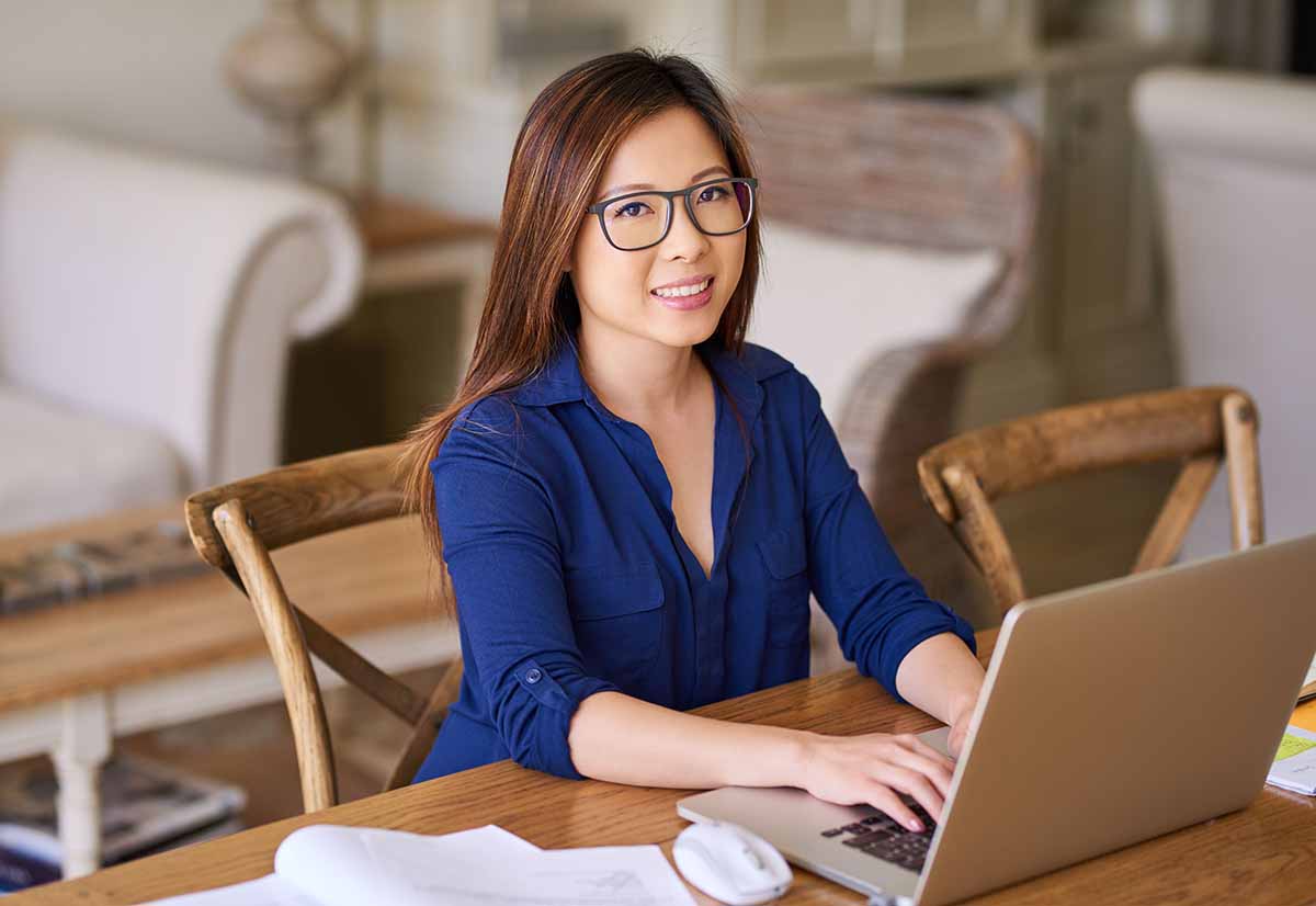 A smiling Asian woman is working from home on a laptop.
