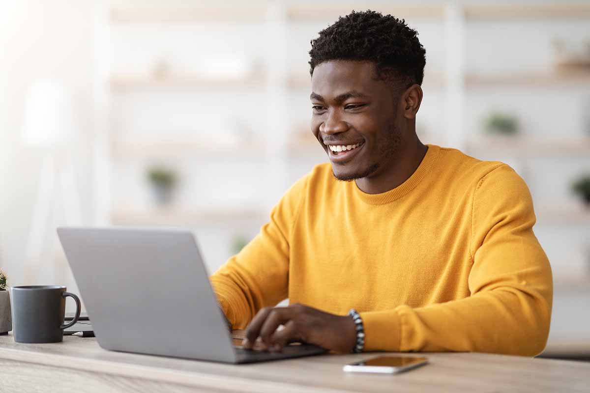 A smiling young African American guy typing on a laptop keyboard while sitting at a desk.