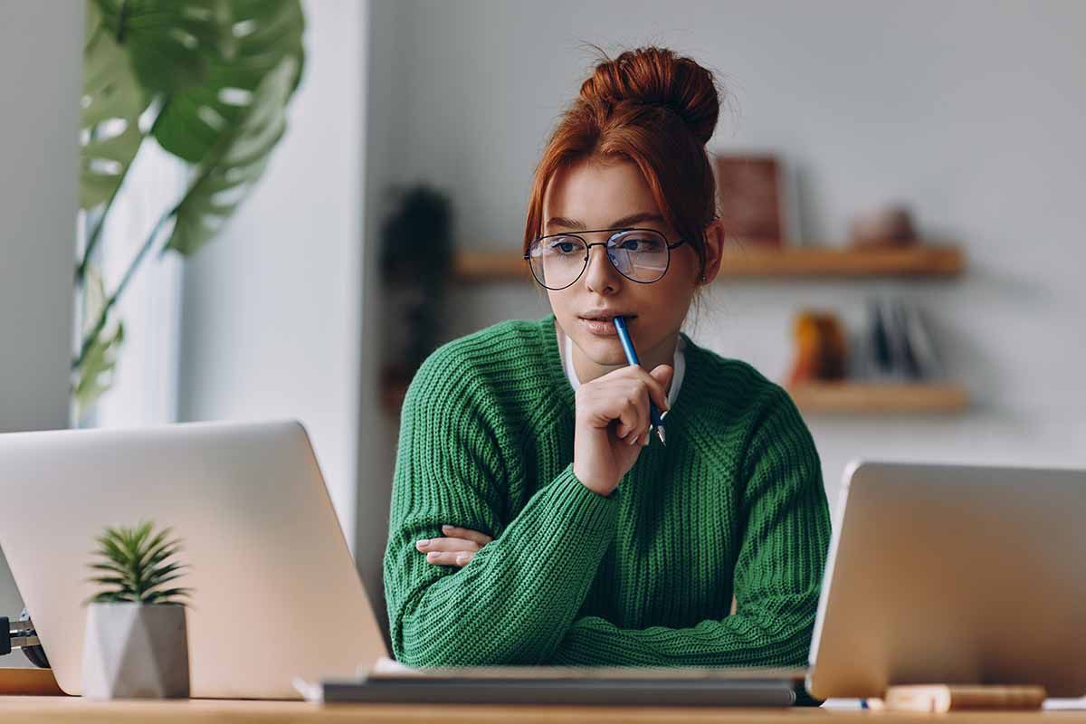 A young girl wearing glasses biting on her pen with a puzzled look while looking at her computer screen.