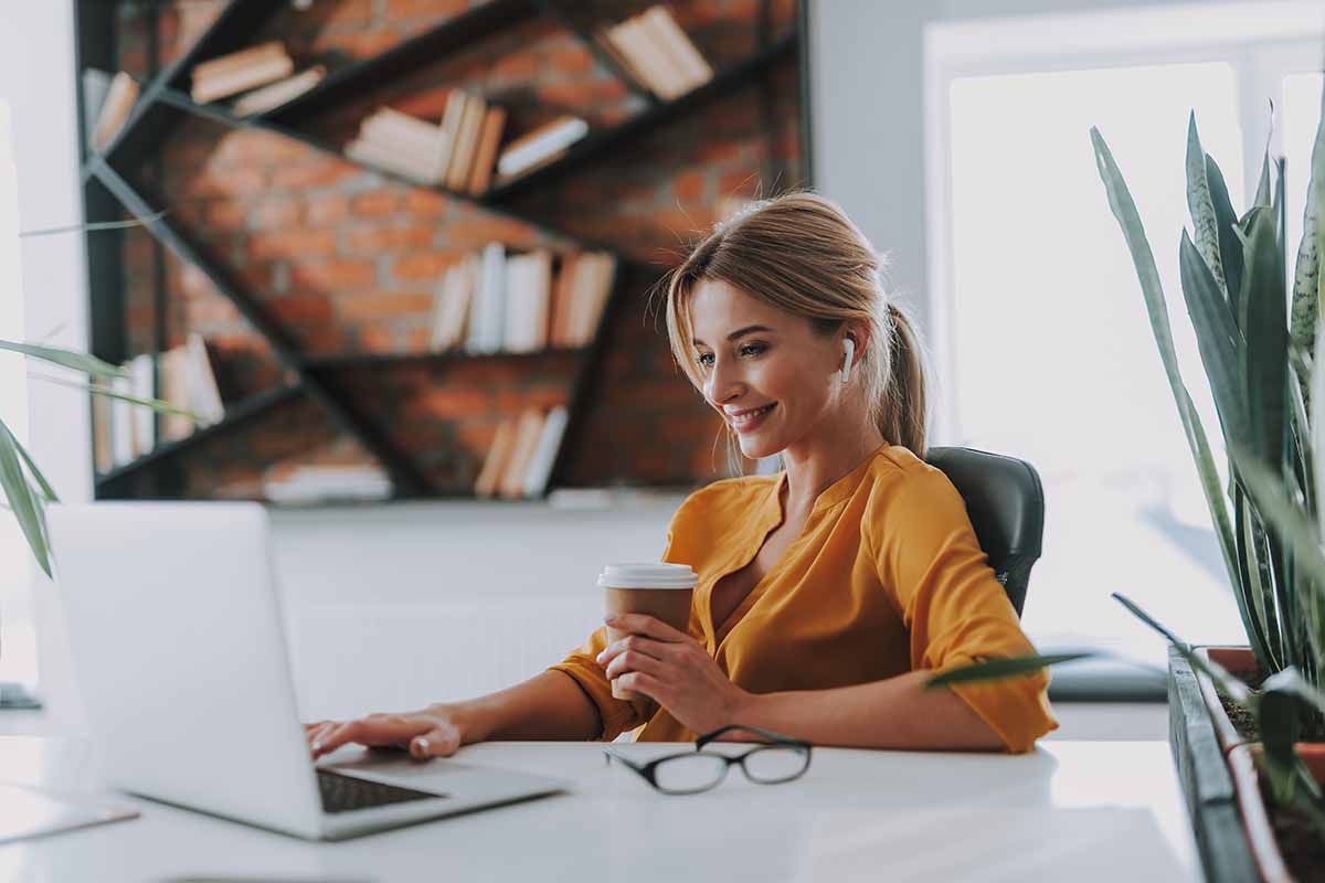A smiling young lady with ear buds in her ears, holding a coffee, and working on a laptop.