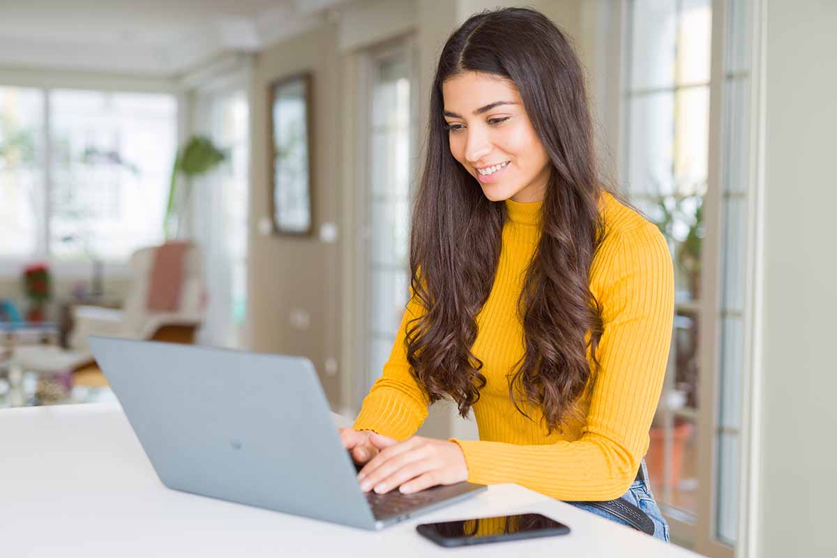 A smiling young lady in a kitchen working on a laptop with her cellphone next to the laptop.