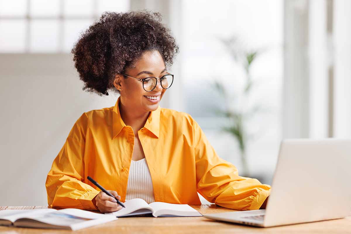A young smiling African American woman working on a laptop while writing in a notebook.
