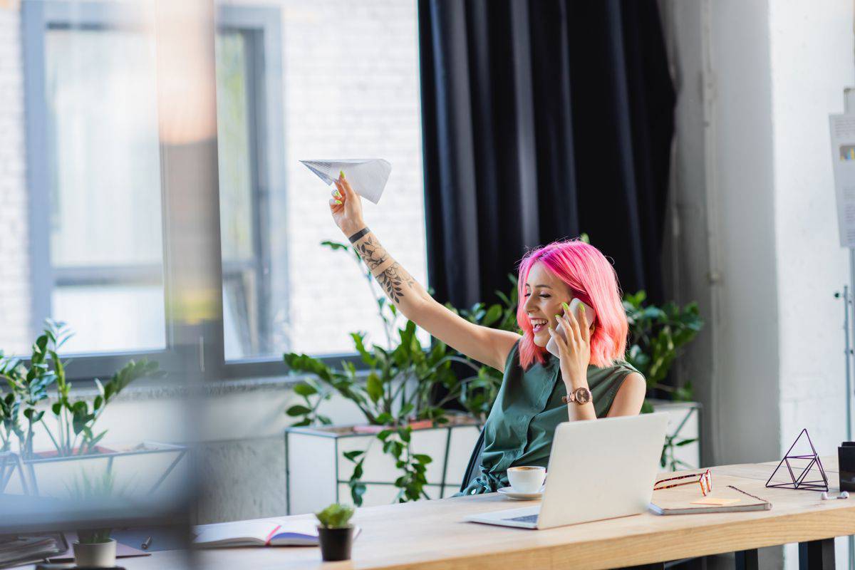 Happy businesswoman with pink hair talking on smartphone while throwing paper plane.